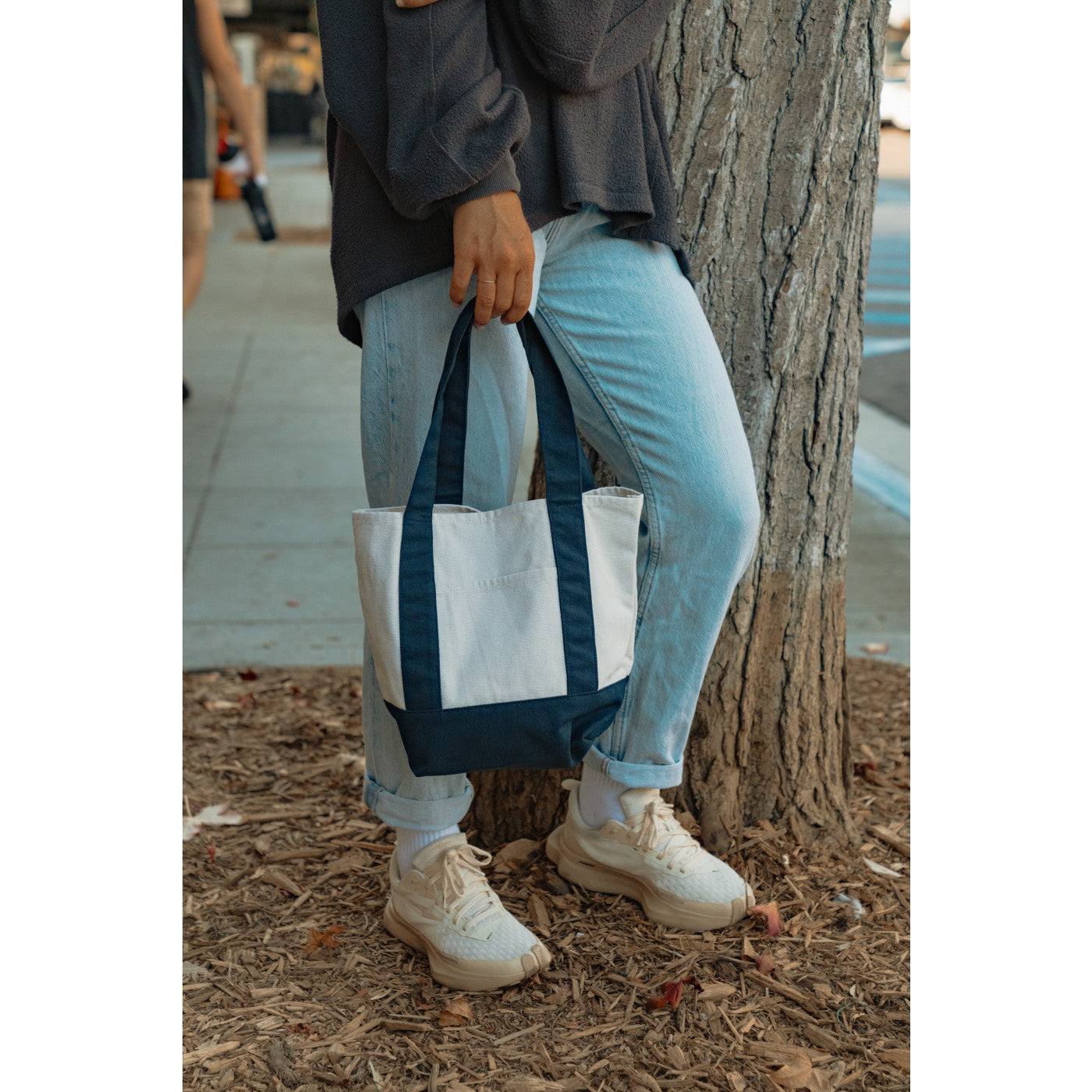 Person holding a tote bag by a tree on a sidewalk