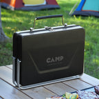 Black portable grill with 'CAMP' branding on a wooden table outdoors, with tents in the background.