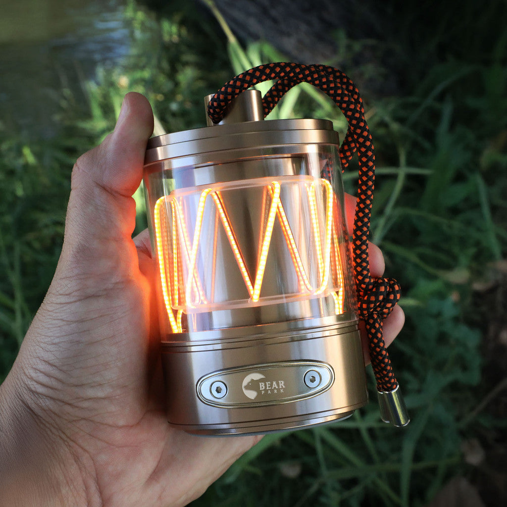 Hand holding a small, illuminated camping lantern with a rope handle against a green foliage background.
