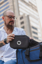 A man in a white shirt and glasses holds a Nomad Must Haves Renew Tech Case with a circular white logo, next to a blue duffel bag, with blurred skyscrapers in the background.