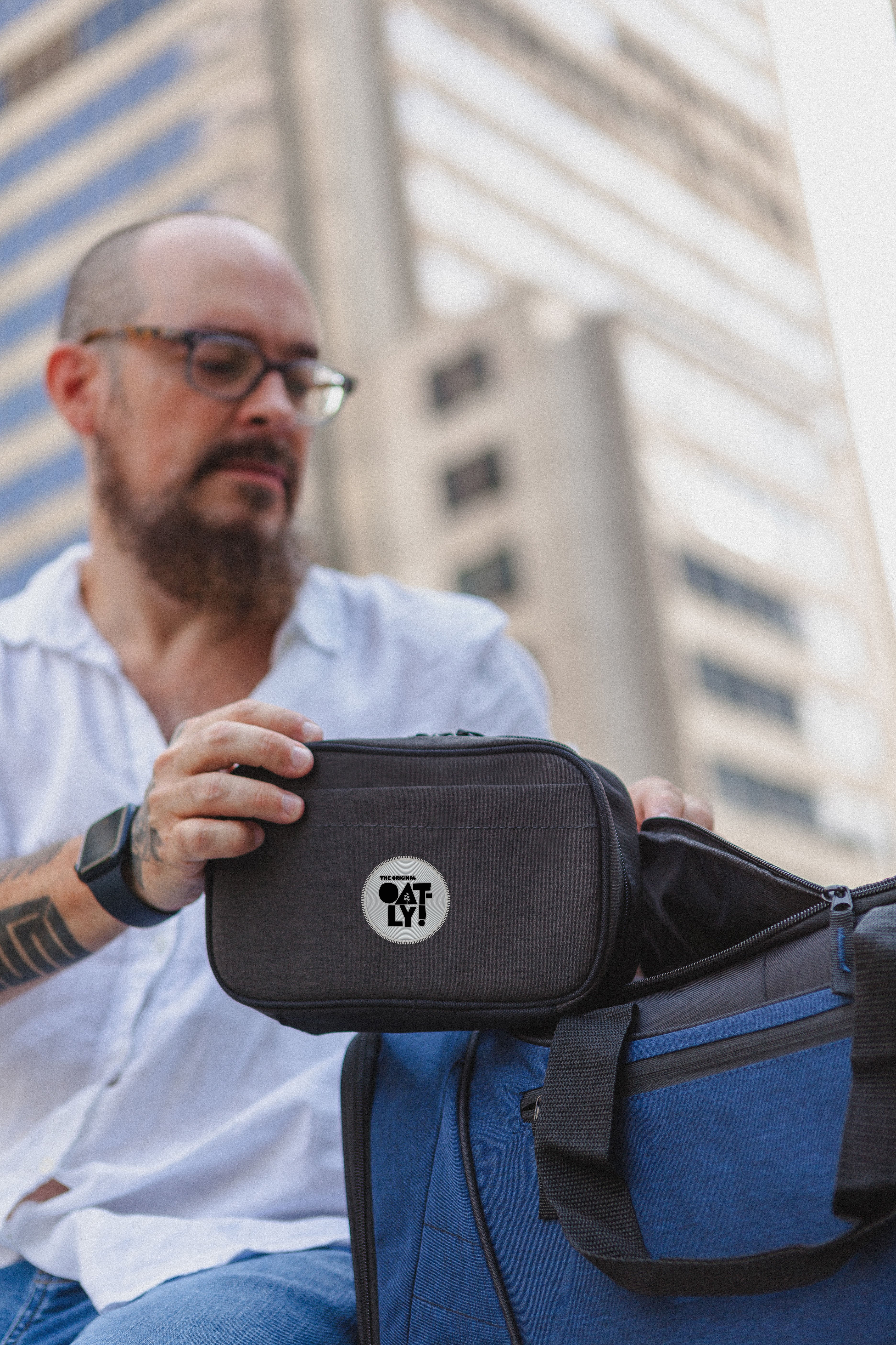 A man in a white shirt and glasses holds a Nomad Must Haves Renew Tech Case with a circular white logo, next to a blue duffel bag, with blurred skyscrapers in the background.