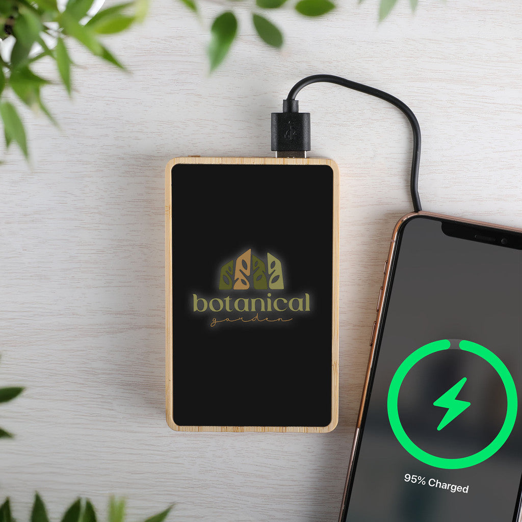 Portable power bank with  logo charging a smartphone on a light surface with greenery.