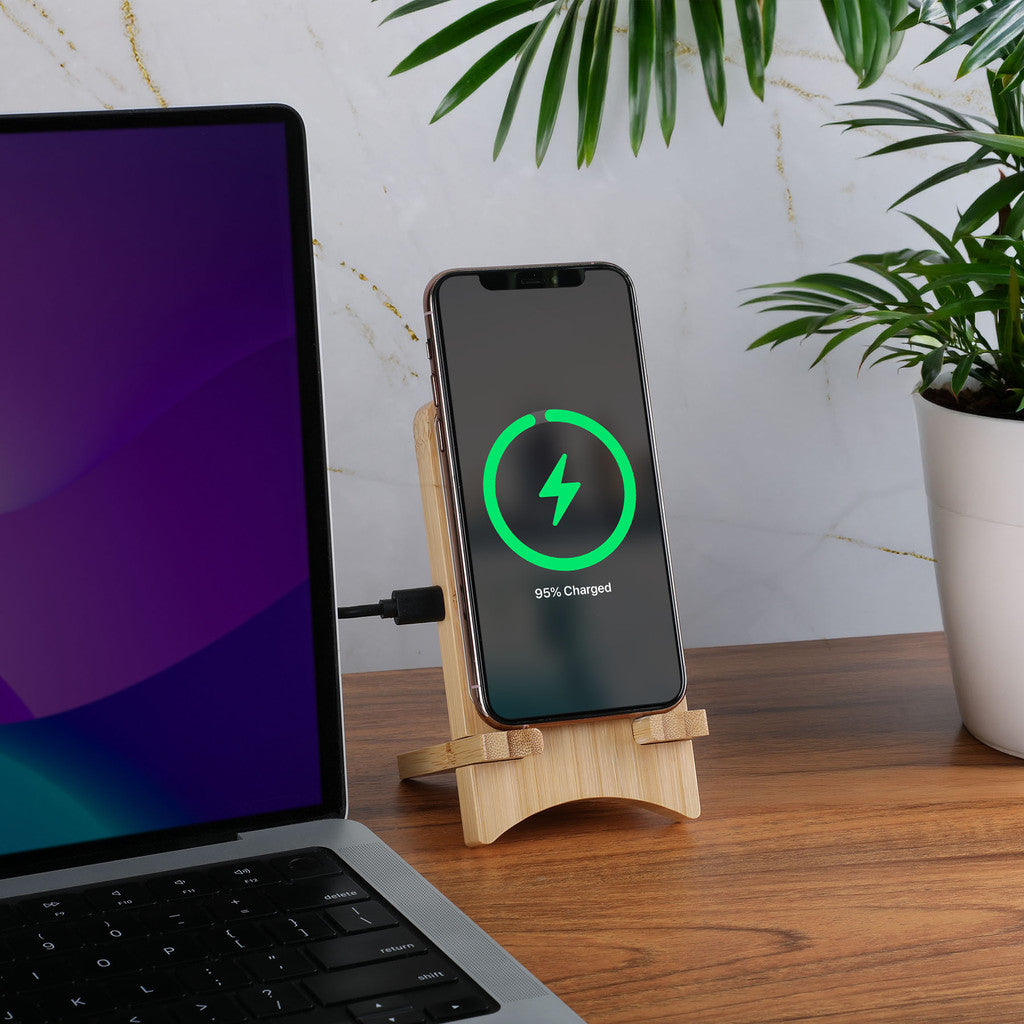 Smartphone on a wooden stand charging with a laptop and plant in the background