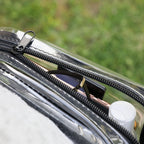 Close-up of a clear zippered pouch with a black zipper and black strap, shown outdoors with a blurred green background. Contents inside the pouch are partially visible, including a dark rectangular item and a white cap.