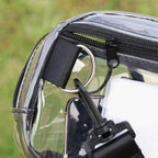 Close-up of a transparent waterproof bag with black zippers, a black webbing strap, a silver metal D-ring, and a black clip, shown outdoors with blurred green grass in the background.
