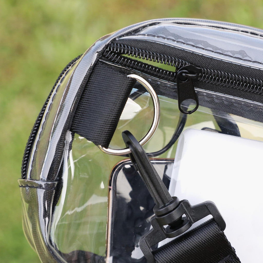 Close-up of a transparent waterproof bag with black zippers, a black webbing strap, a silver metal D-ring, and a black clip, shown outdoors with blurred green grass in the background.