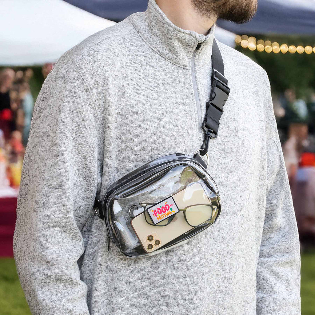 A man wearing a gray quarter-zip sweatshirt carries a Clear Crossbody Sports Pack with a black strap and zipper. The pouch contains a beige smartphone, eyeglasses, and a small item with a colorful 'FOOD' logo.
