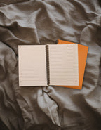 An open lined spiral notebook with white pages lies on top of an orange notebook , placed on rumpled gray fabric.