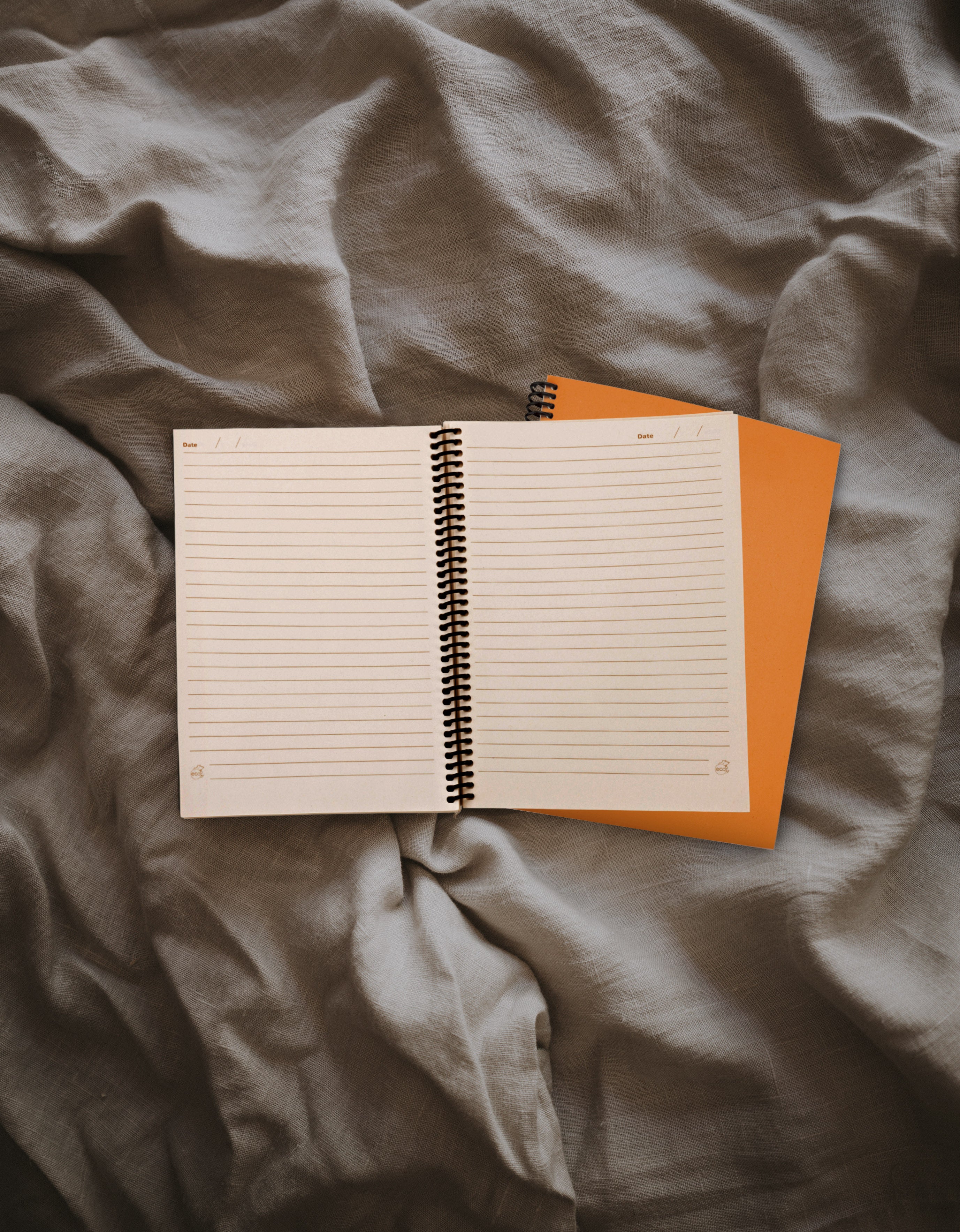 An open lined spiral notebook with white pages lies on top of an orange notebook , placed on rumpled gray fabric.