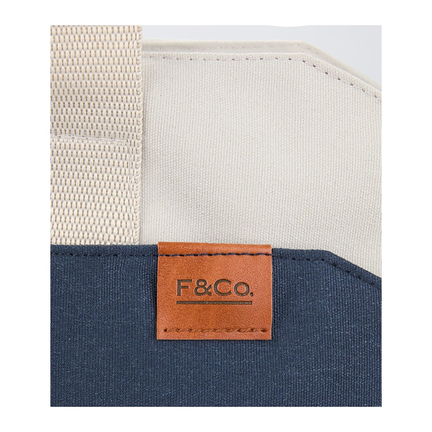 Close-up of a blue bag with a brown leather tag featuring 'F&Co.' on a white background.