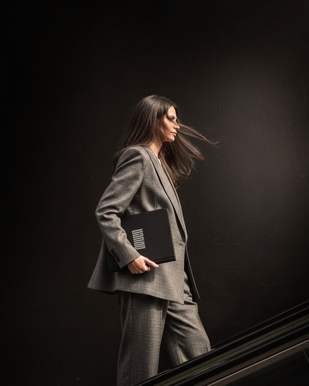 A woman in a tailored gray blazer and matching trousers walks down stairs while holding a MODQ A4 Portfolio against a dark studio background.