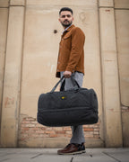 A man wearing a brown corduroy jacket, gray jeans, and brown leather shoes carries a large black duffel bag with a yellow logo patch, standing against a beige brick wall.