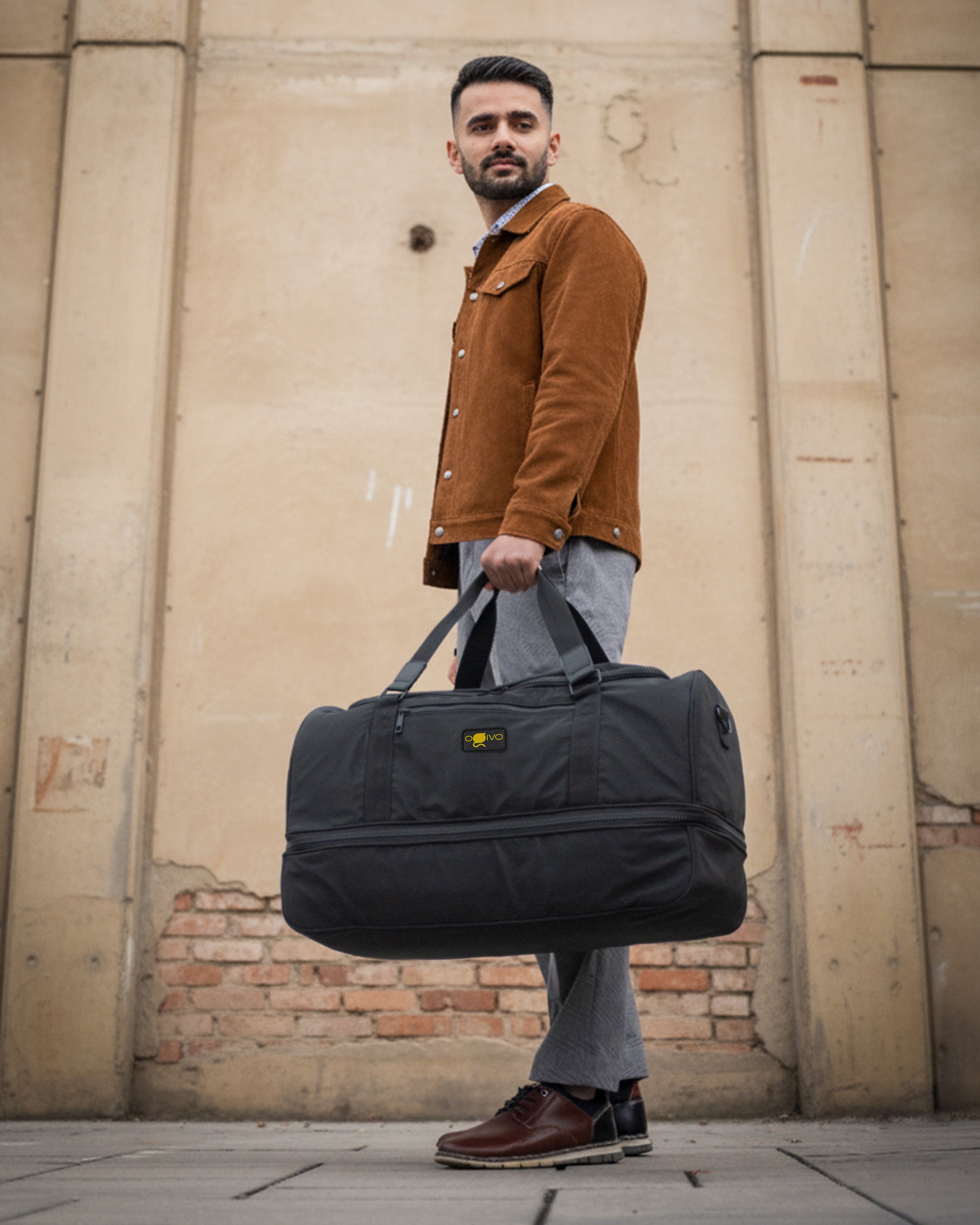 A man wearing a brown corduroy jacket, gray jeans, and brown leather shoes carries a large black duffel bag with a yellow logo patch, standing against a beige brick wall.